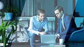 Ein Mitarbeiter und ein Interessent halten in einem Büro ein Verkaufsgespräch. Der Mitarbeiter zeigt dem Interessenten etwas auf einem Laptop. 