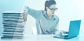 Das Bild zeigt eine Frau, die einen Papierstapel wegschiebt und am PC arbeitet.