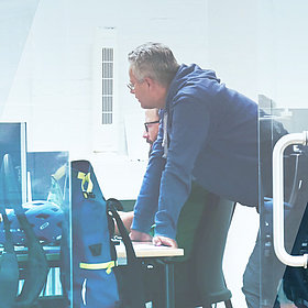 Das Bild zeigt zwei business//acts Mitarbeiter, einer im Hintergrund daneben sitzend an einem Schreibtisch vor mehreren Monitoren, der andere stehend und sich mit den Händen auf dem Schreibtisch abstützend