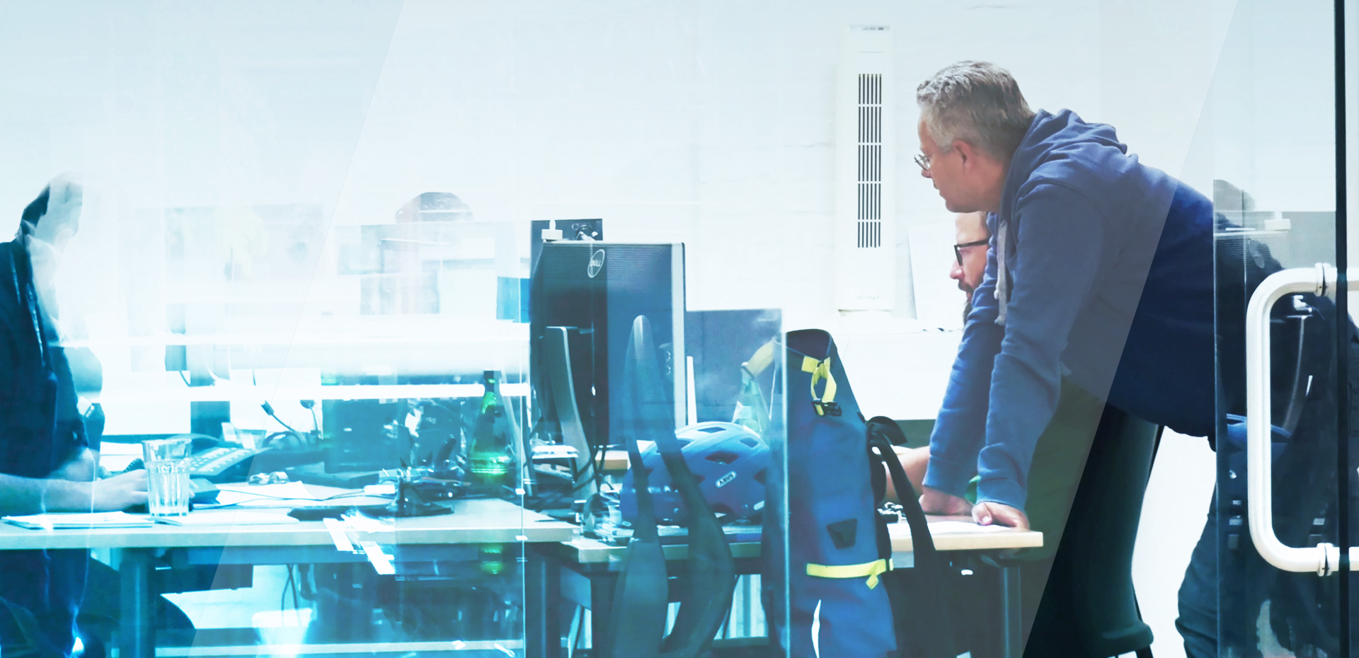 Das Bild zeigt zwei business//acts Mitarbeiter, einer im Hintergrund daneben sitzend an einem Schreibtisch vor mehreren Monitoren, der andere stehend und sich mit den Händen auf dem Schreibtisch abstützend