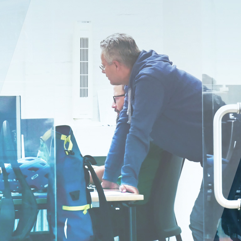 Das Bild zeigt zwei business//acts Mitarbeiter, einer im Hintergrund daneben sitzend an einem Schreibtisch vor mehreren Monitoren, der andere stehend und sich mit den Händen auf dem Schreibtisch abstützend
